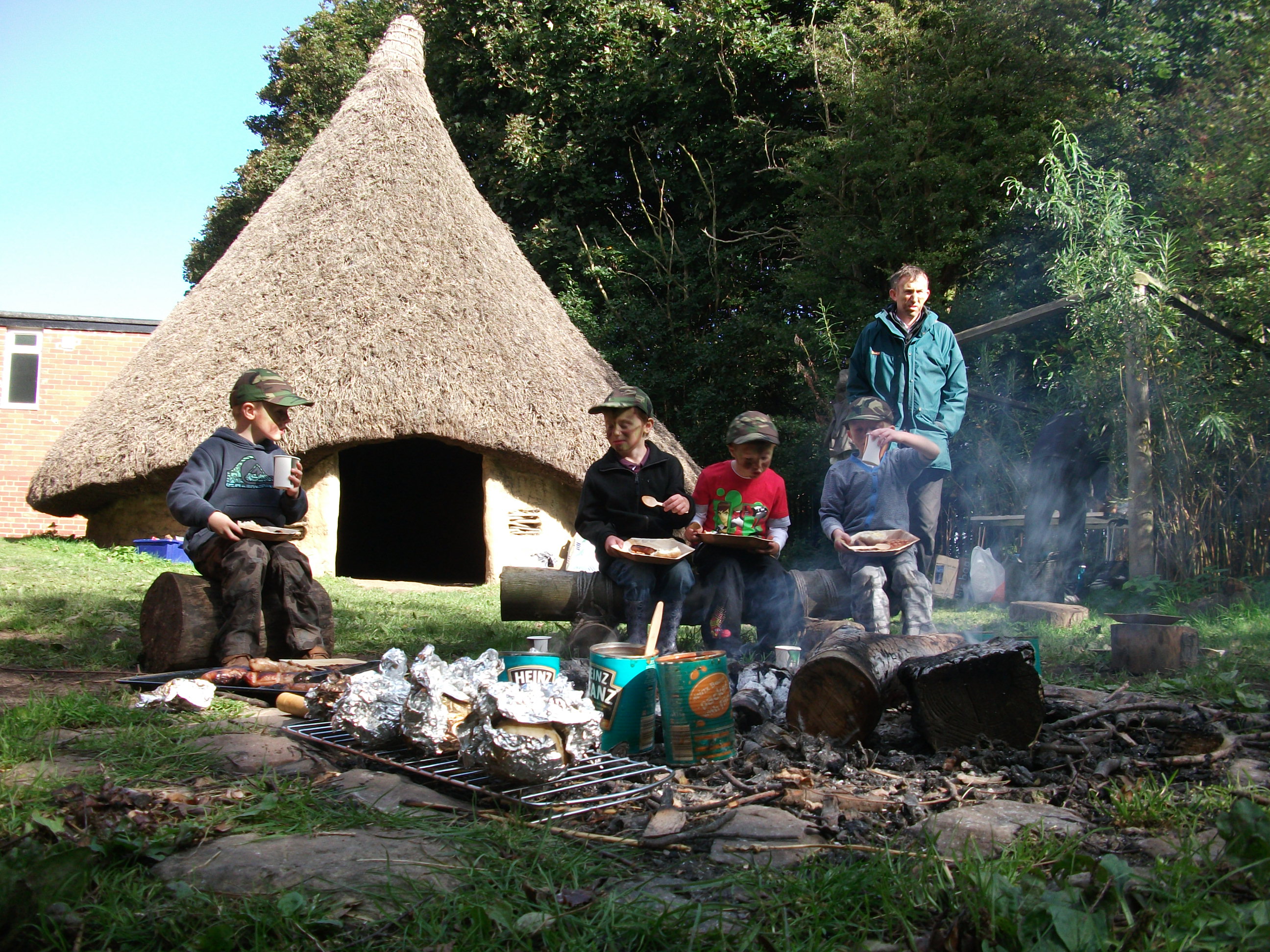 Children and an adult around an outdoor fire in front of an old thatched dwelling