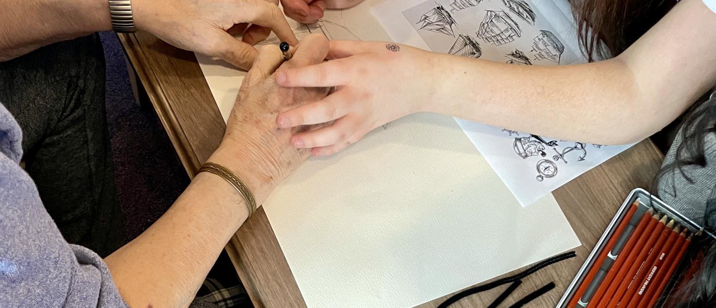 Young person's hands assisting older hands to draw a ship using a pencil and paper