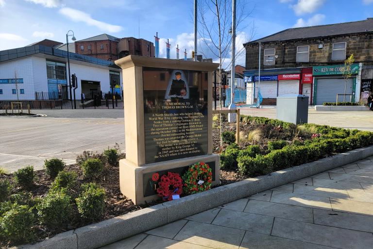 A stone memorial to Thomas brown with an inscription and a row of shops in the background