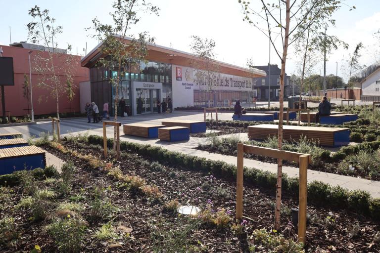 The town square with flower beds, trees and seats and some people sitting and walking around