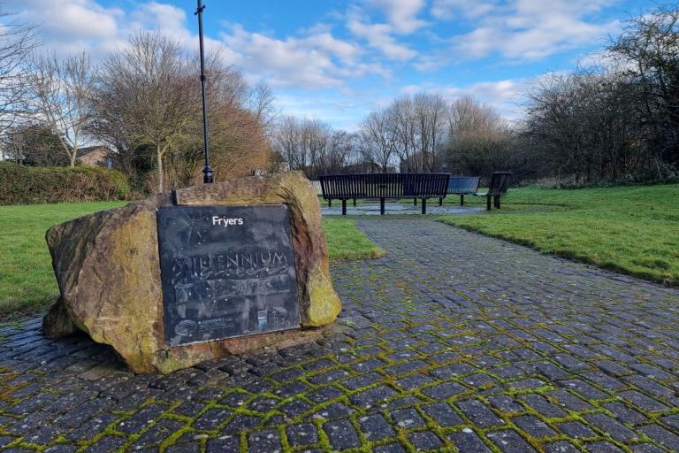 A colour photo of a plaque fixed to a stone saying Fryers Millennium Green with paving leading towards a row of metal park benches with trees surrounding a grassed area, blue skies with white clouds