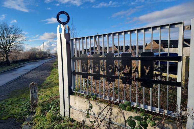 A metal gate with the word Waggonways in wrought iron lettering with a bridle path leading off into the distance with trees and bushes, wintery but blue skies
