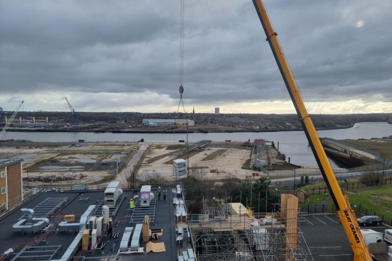 A colour photo of a crane lowering new aircon units on to the roof of Segedunum with the River Tyne behind
