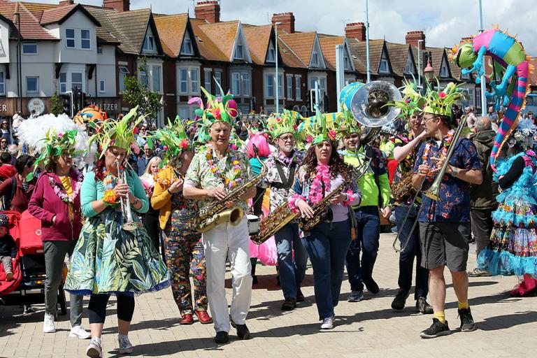 A colour photo of musicians with saxophones and trombones in carnival clothing walking along the street in Whitley Bay 