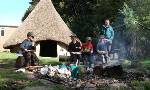 Children and an adult around an outdoor fire in front of an old thatched dwelling