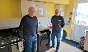 Two men standing in a bright kitchen with a tumble dryer between them